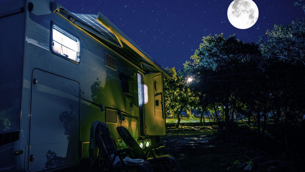 A parked RV at night with its interior lights on, two chairs outside, trees surrounding the area, and a full moon in the starry sky.