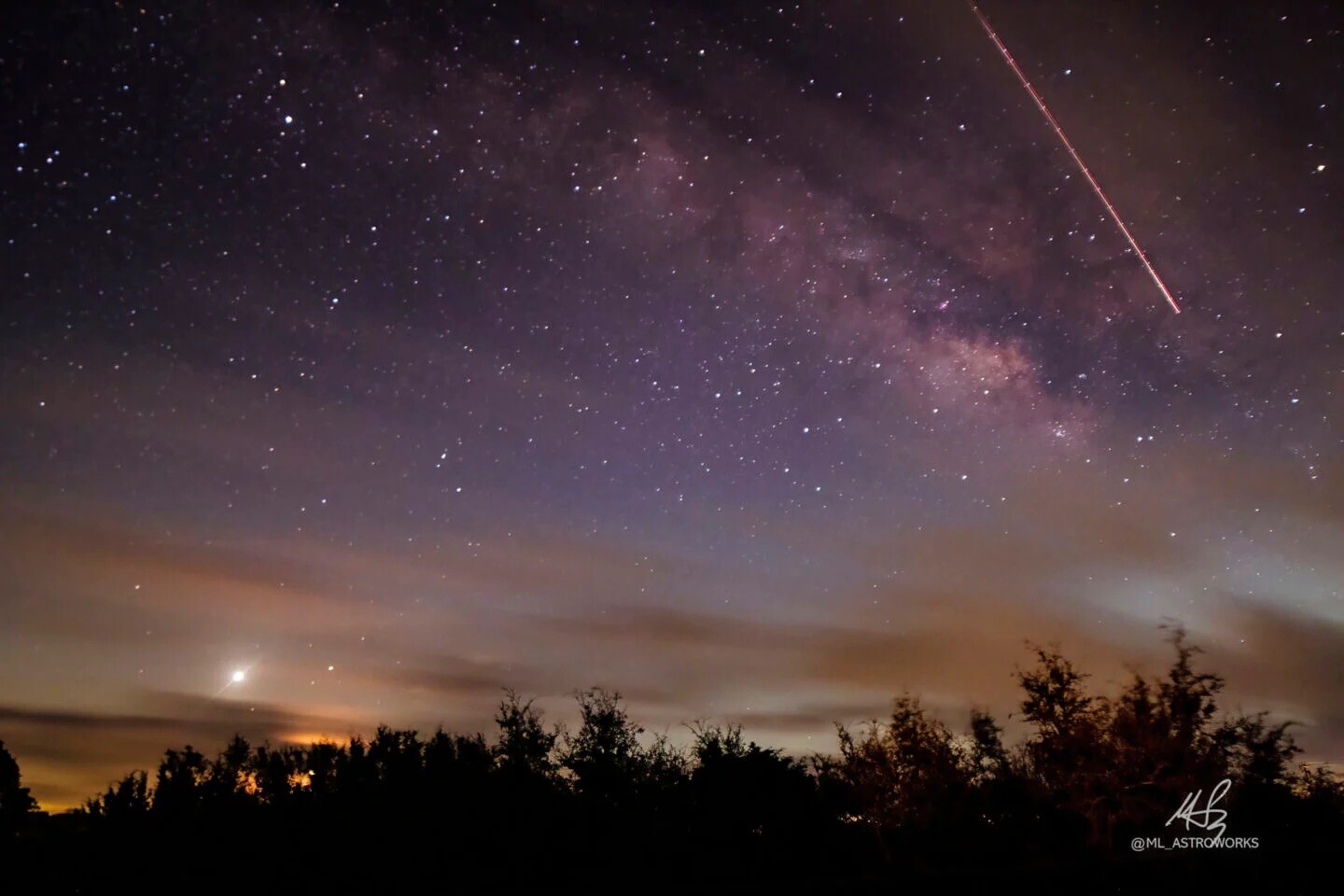 Starry night sky with Milky Way visible, streaking light across the top right, silhouetted trees at the bottom, and a bright object near the horizon.