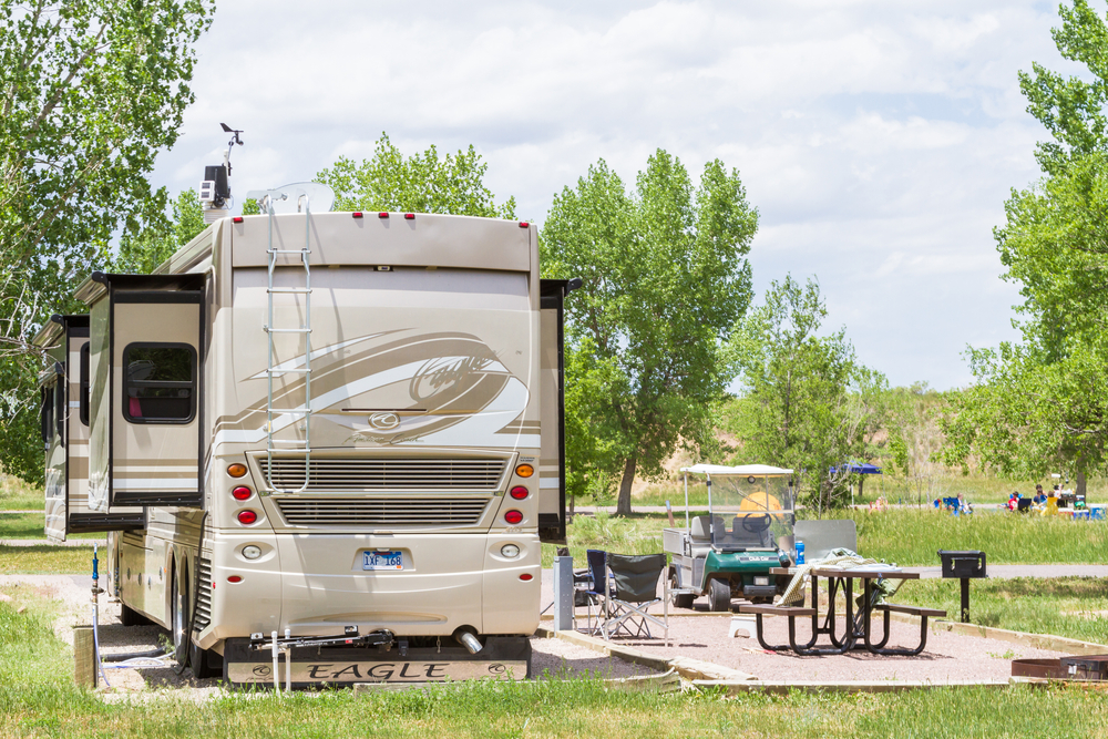 A beige RV is parked at a campsite with a golf cart, folding chairs, and a picnic table. Trees and greenery surround the area under a partly cloudy sky.