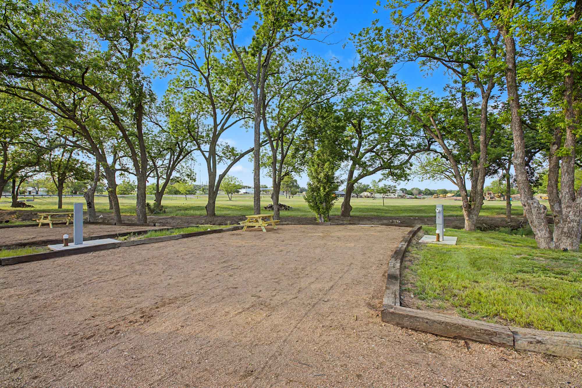 A gravel RV site with utility hookups, picnic tables, and surrounding green trees under a clear blue sky.