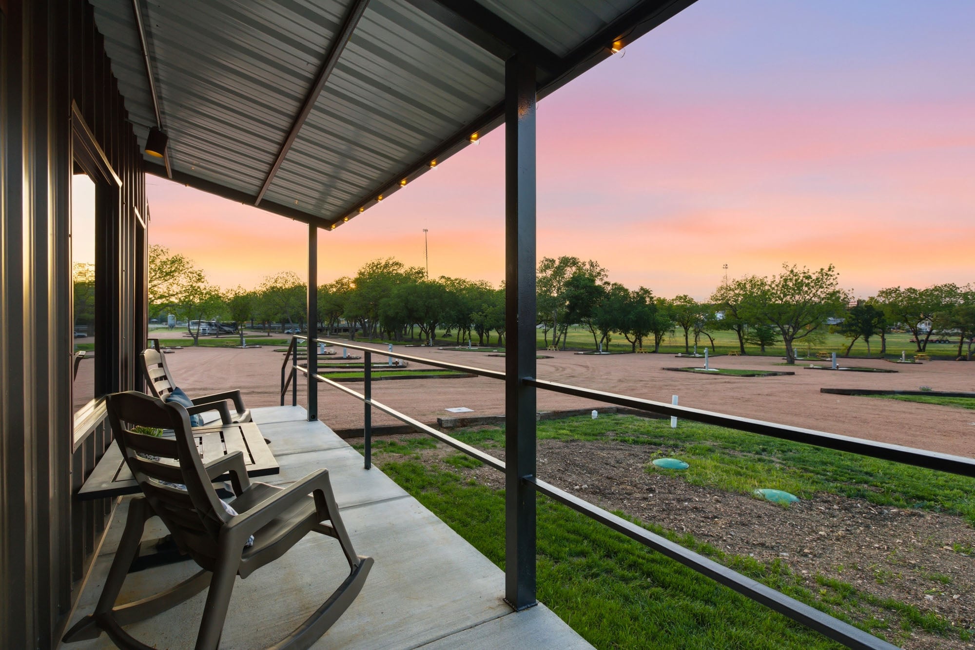 Covered porch with two rocking chairs overlooking a gravel parking area, grassy lawn, and trees at sunset.