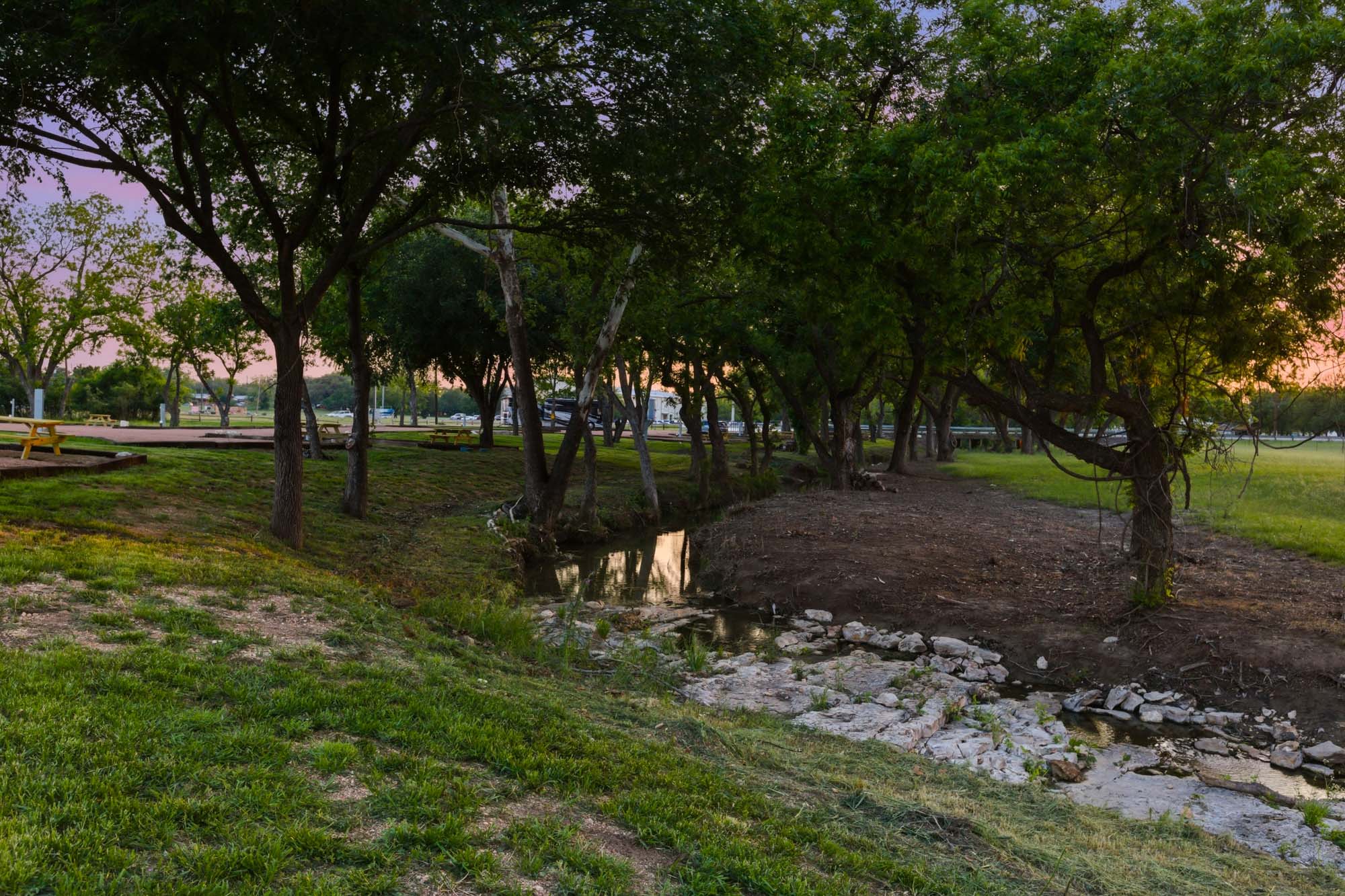 A small creek runs through a grassy park area with trees on both sides, rocks in the creek bed, and benches visible in the background at sunset.
