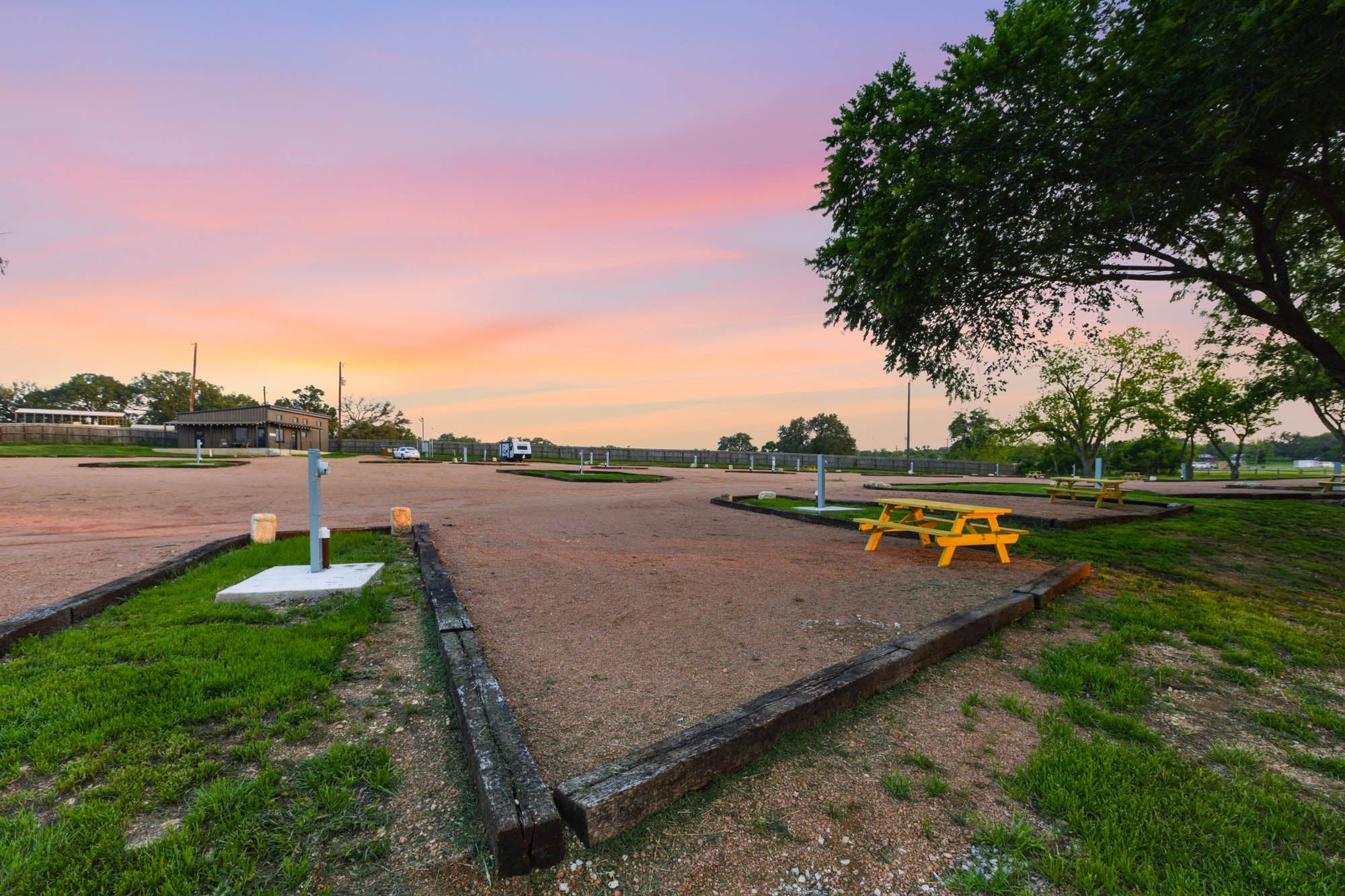 Empty RV parking spaces with utility hookups and a yellow picnic table are shown at sunset, with trees and a building in the background.