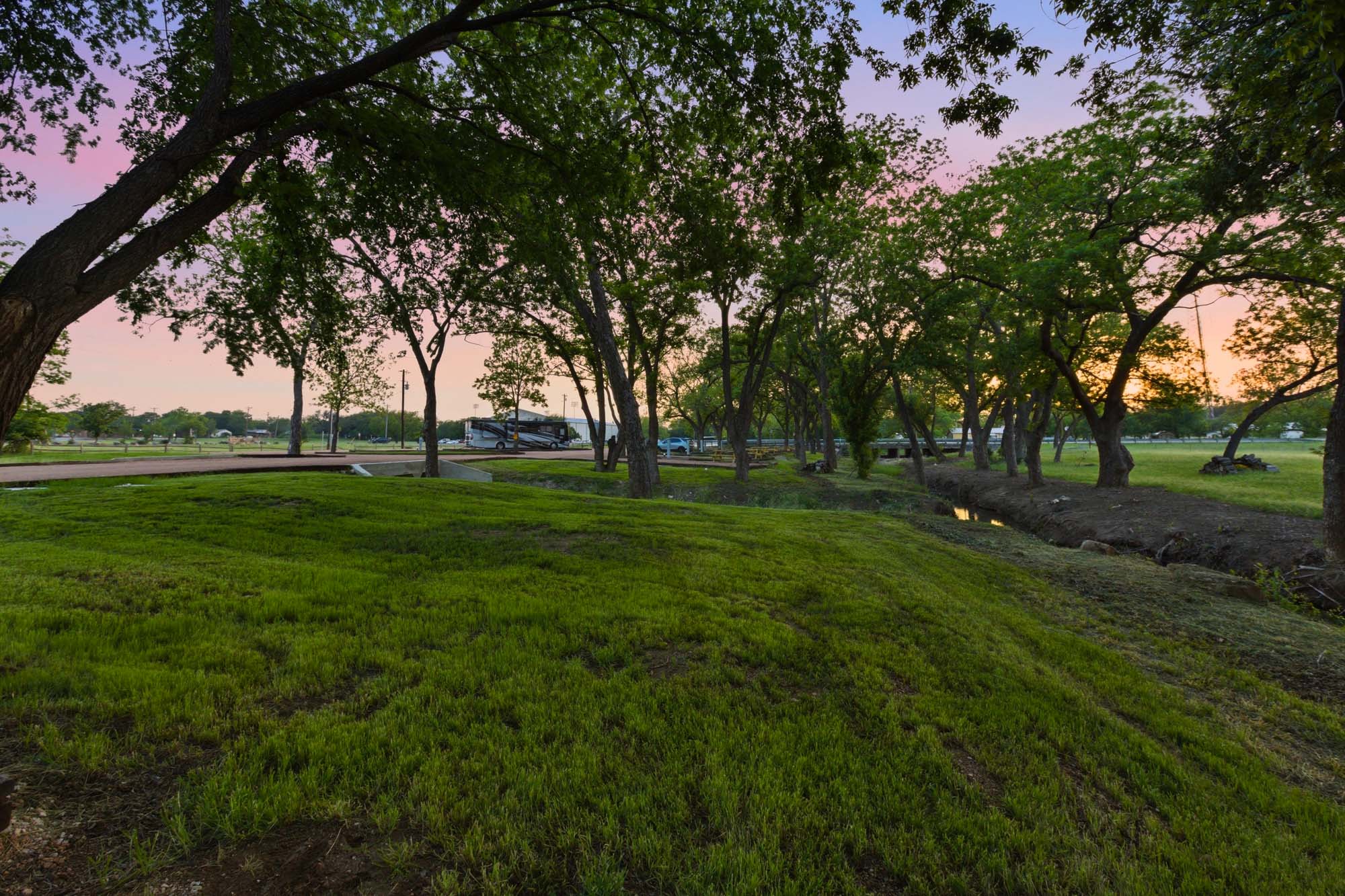 A grassy park area with scattered trees at sunset, a paved path to the left, and an RV parked in the background near a small creek.