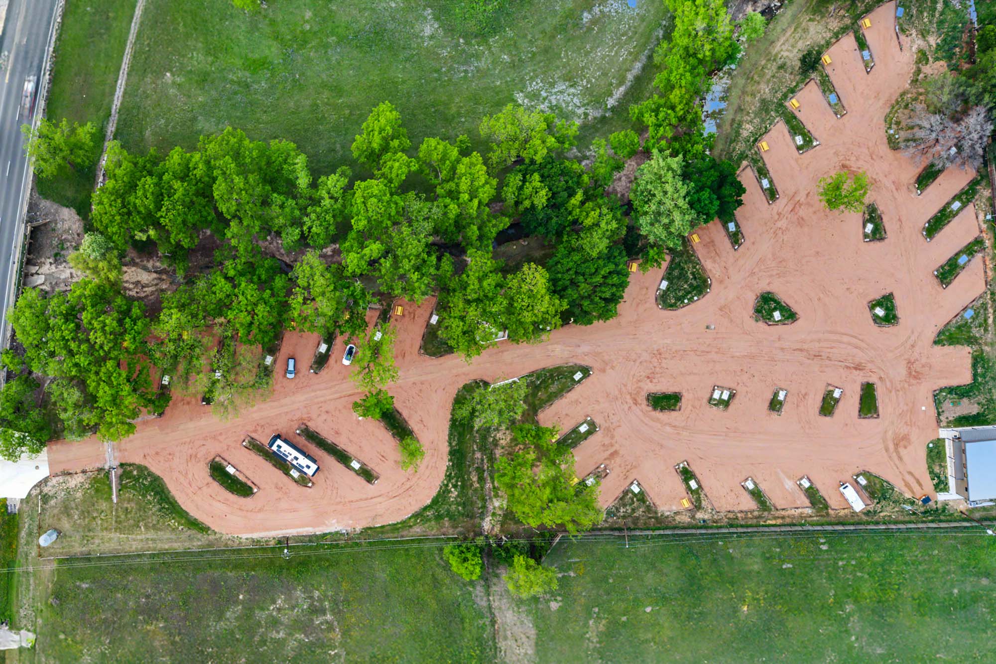 Aerial view of a campground with dirt roads, parking spots, scattered vehicles, and surrounding green trees and grass.