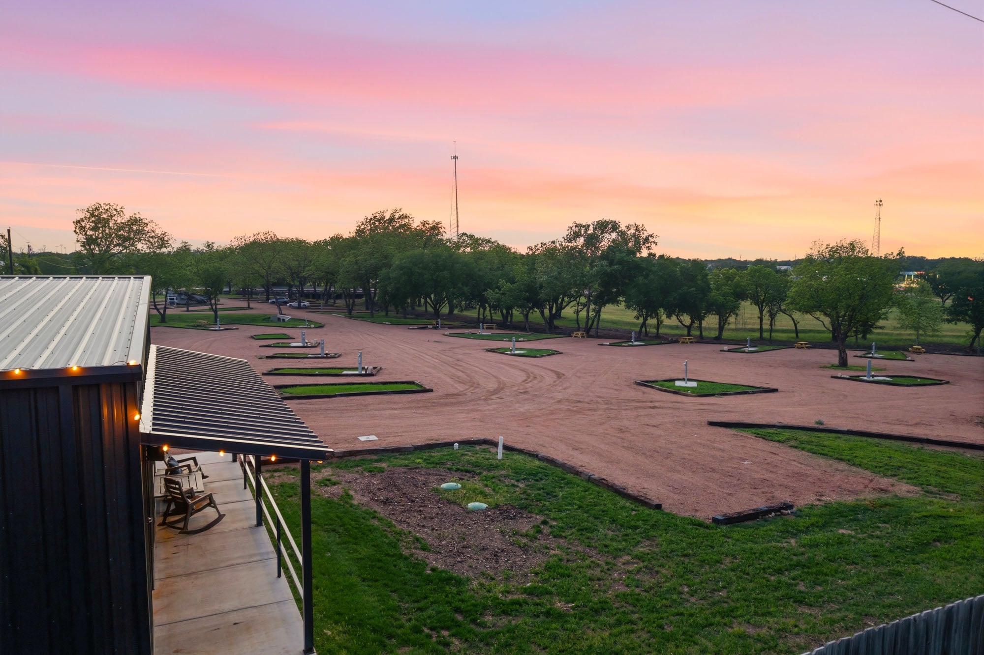 A gravel parking area with marked spaces for RVs or trailers, bordered by grassy plots and trees, is shown at sunset next to a covered porch with chairs.