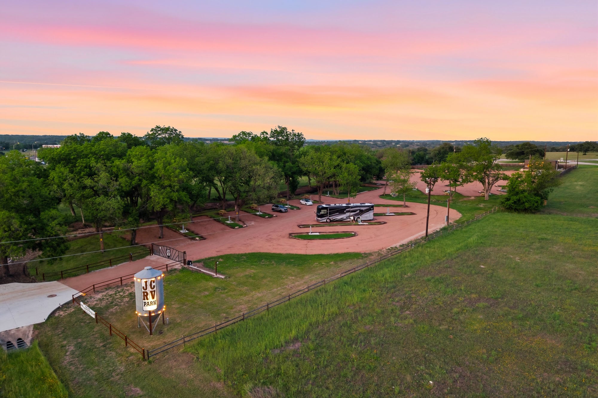 Aerial view of an RV park with several empty lots, one RV parked, surrounding trees, and a colorful sunset sky in the background.